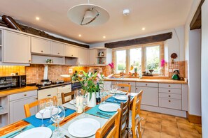 A kitchen with white cabinets, a dining table set for four, and a window with a view of greenery. - Dunford Barn (Lechlade)