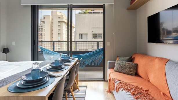A modern hotel room with a balcony, a dining area with blue plates, a sofa with a throw blanket, and a flat-screen TV mounted on the wall. - StayHome Frei Caneca Rooftop Gym Jacuzzi (São Paulo)