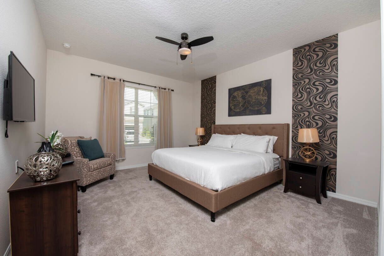 A bedroom with a bed, a desk, a chair, a nightstand, a ceiling fan, a window with curtains, and a wall with a patterned wallpaper.