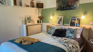 Ein ordentlich bezogenes Bett mit blauer Decke und Kissen, ein Nachttisch mit Lampe sowie Kunstwerke an den Wänden.