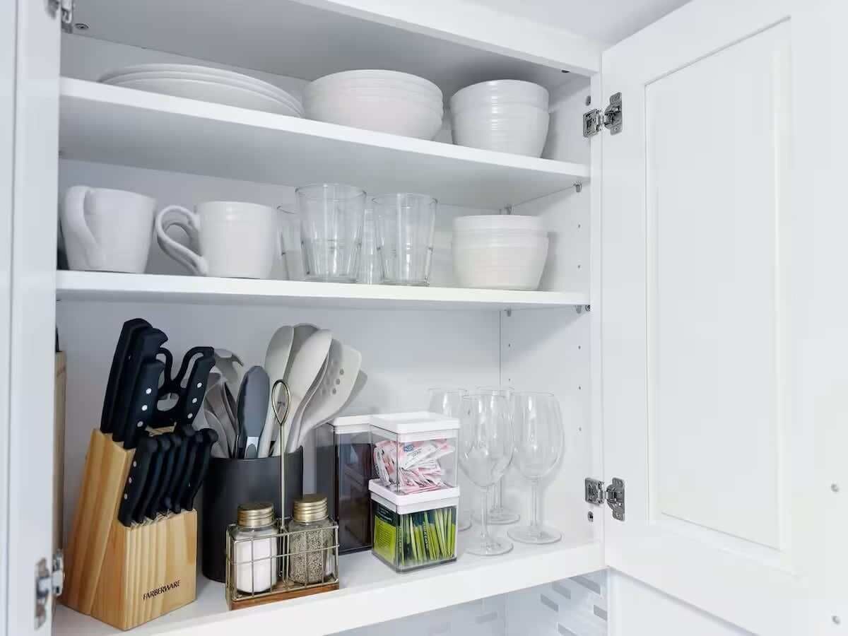 Un armario de cocina con estantes blancos que contiene platos, tazas y vasos.