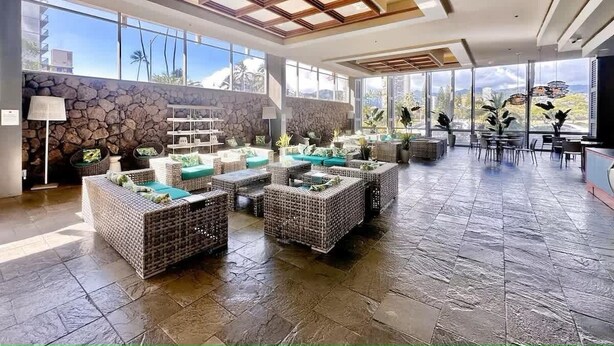 A spacious lobby with wicker furniture, stone walls, and large windows offering views of palm trees and buildings. - @ Marbella Lane- Trendy Waikiki Oasis Near Beach (Honolulu)