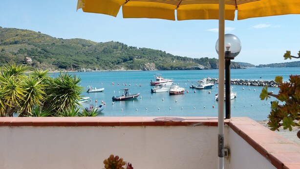 Un balcone con un ombrellone giallo, affacciato su una marina con barche, vegetazione e un paesaggio collinare.