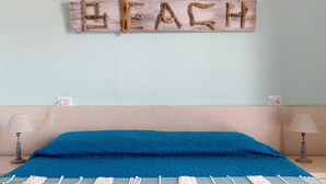 Un letto con biancheria azzurra, un cartello di legno con la scritta 'BEACH' sulla parete e due abat-jour.