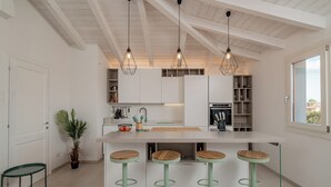 A modern kitchen with white cabinets, a central island with stools, pendant lights, and a view through a window. - VILLA CIELO DI TAVOLARA, just a few steps from the sea with breathtaking views. (Porto San Paolo)