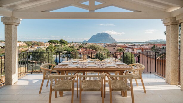 A covered outdoor dining area with a table set for six, surrounded by wicker chairs, overlooking a coastal town with a prominent mountain in the distance. - VILLA CIELO DI TAVOLARA, just a few steps from the sea with breathtaking views. (Porto San Paolo)