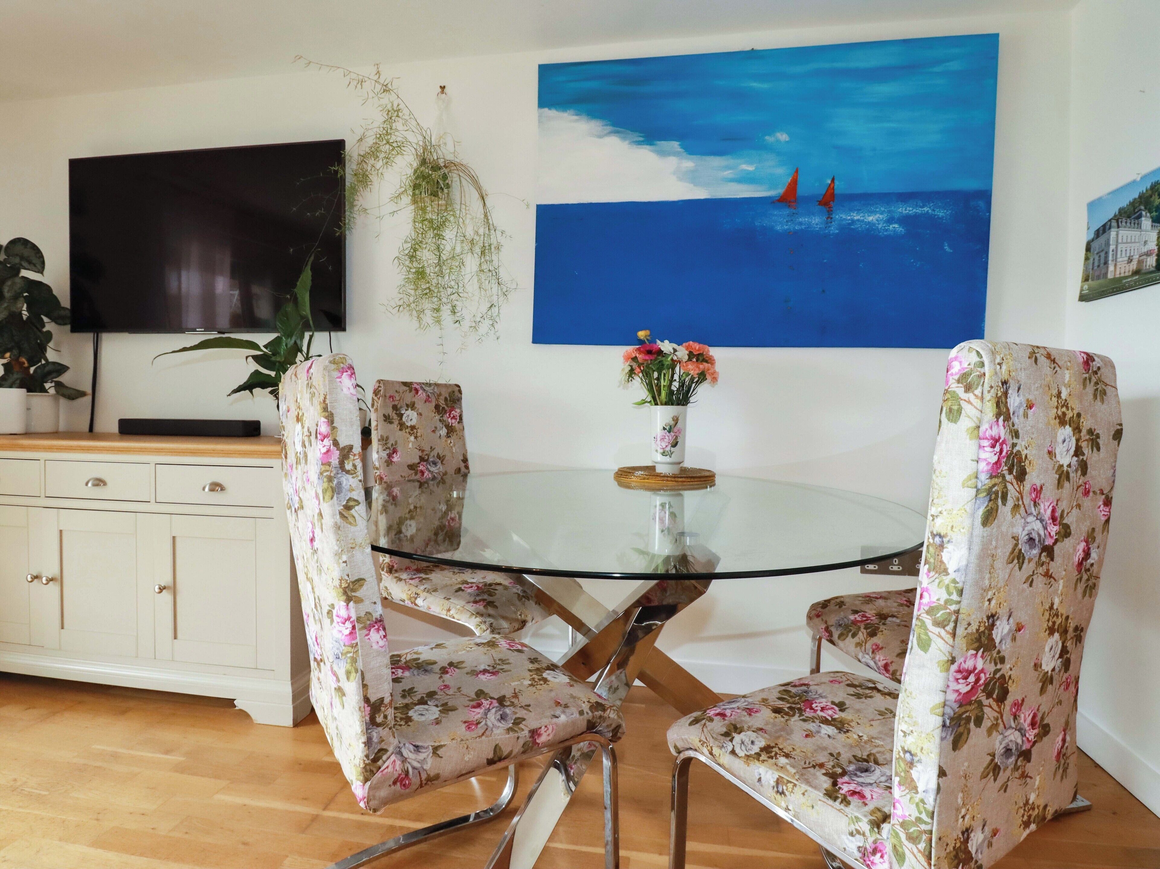 A dining area with a glass table surrounded by floral-patterned chairs, a flat-screen TV, and a painting of a seascape.