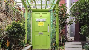 A green door with a 'VACANCIES' sign, a small garden area with plants and a bench, and a brick wall. - Number 6 (Blackpool)