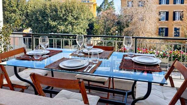 Mesa de vidrio con capacidad para cuatro personas, sillas, barandilla y edificios al fondo.
