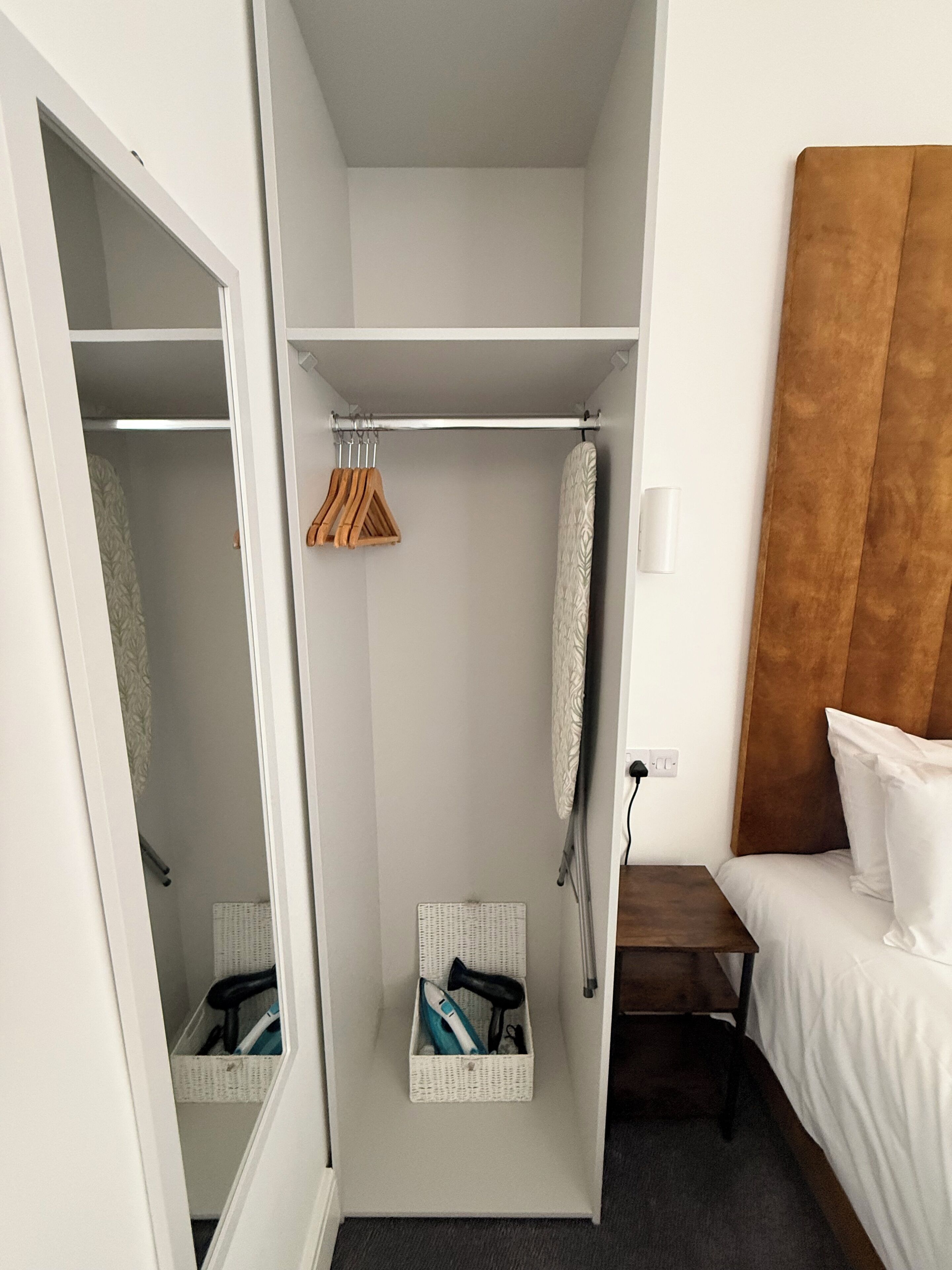 Una habitación de hotel con una cama, cabecera de madera, armario blanco y una pequeña tabla de planchar.