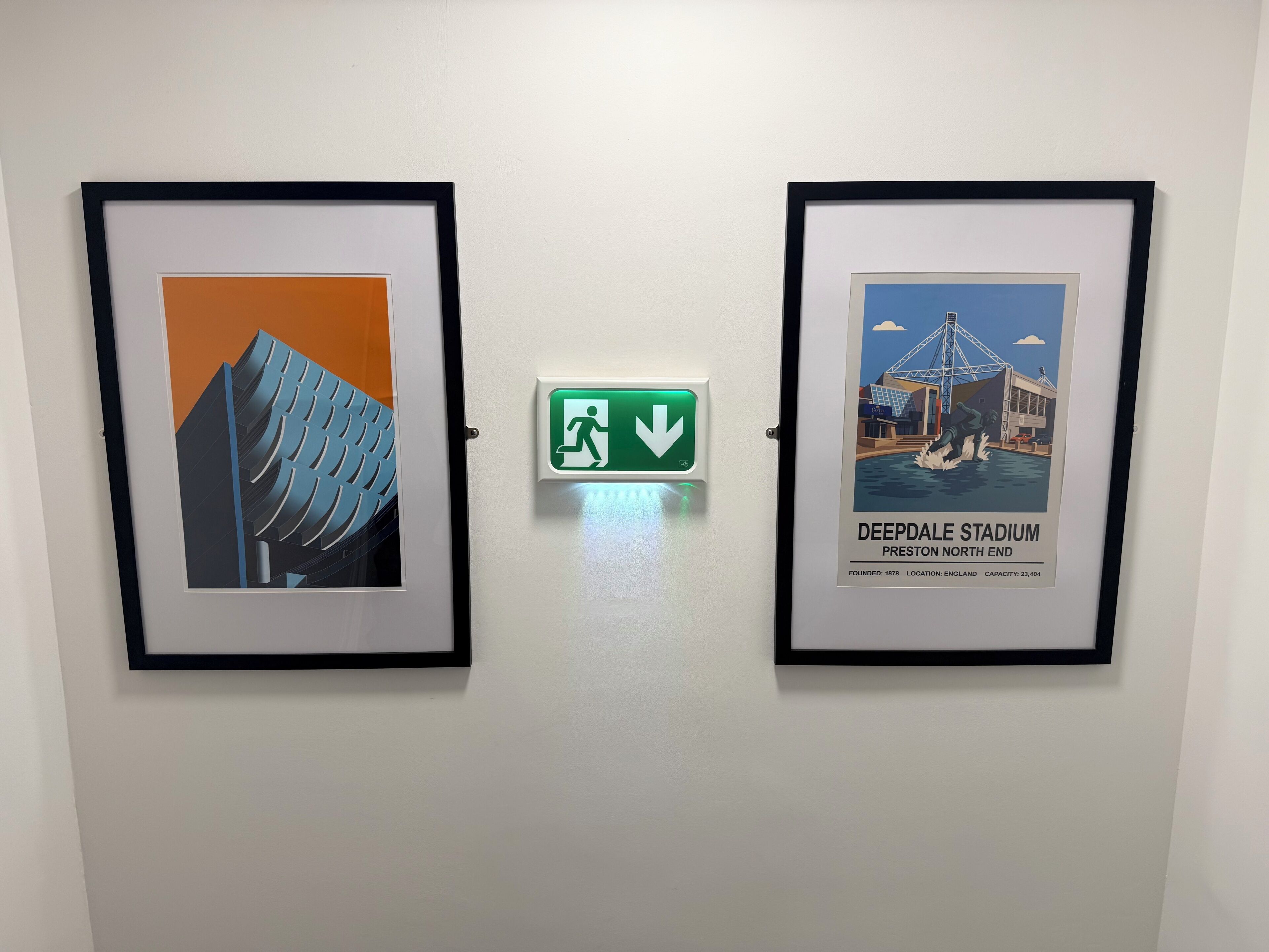 Due opere d'arte incorniciate su una parete, una raffigura un edificio moderno e l'altra un poster di uno stadio.