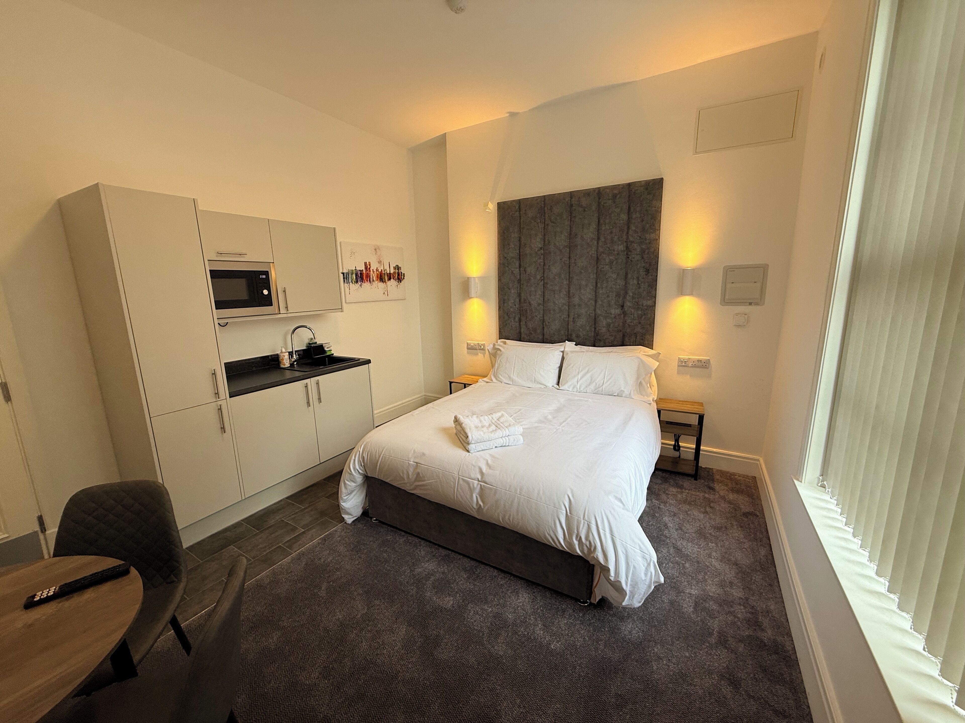 A hotel room with a bed, a desk, a microwave, a sink, and a window with blinds.