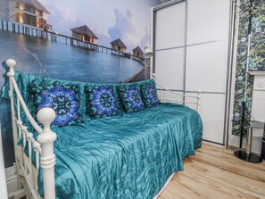 Una cama con colcha verde azulada y almohadas estampadas, cabecera blanca y un mural con bungalows sobre el agua.