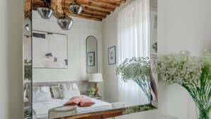 Ein Schlafzimmer mit Bett, Nachttisch, Lampe, Spiegel und einem großen Fenster mit Vorhängen.