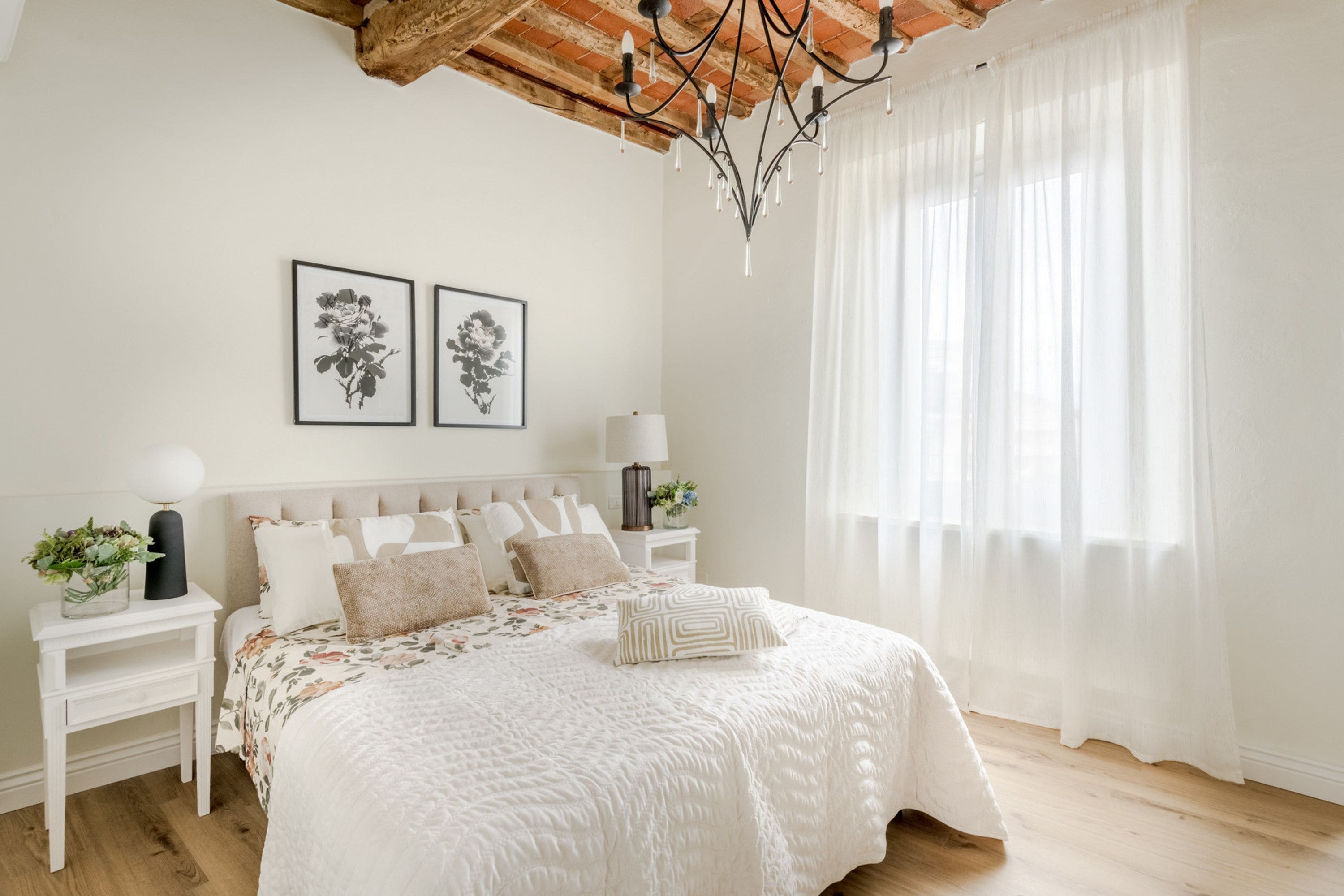 Un dormitorio con una cama, mesitas de noche blancas, una araña y dos cuadros enmarcados con motivos florales en la pared.