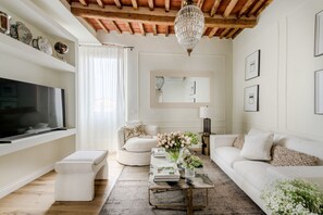 Una sala de estar con televisor, sofás blancos, una mesa de centro y una araña.
