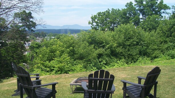 Tre poltrone Adirondack e un tavolo su un prato verde con vista su alberi e montagne lontane.