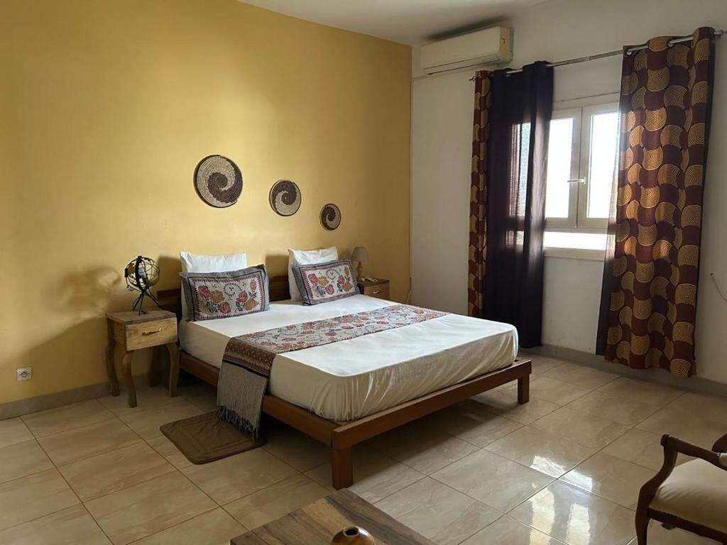 A bedroom with a bed, bedside tables, a nightstand, a window with curtains, and a wall-mounted air conditioner.