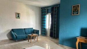 A room with a teal sofa, a small wooden table, a white bed, and a patterned rug. - Jozene House  (Ngaparou)