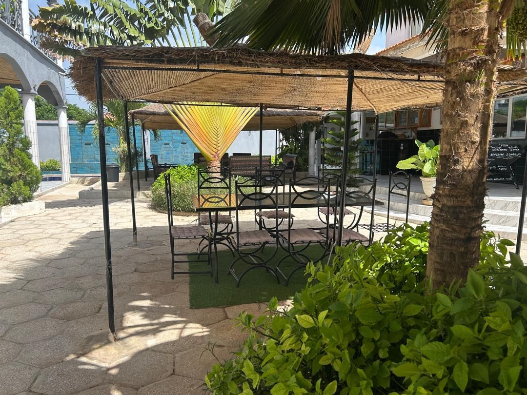 An outdoor seating area with a thatched roof, surrounded by greenery and a palm tree.