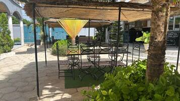 An outdoor seating area with a thatched roof, surrounded by greenery and a palm tree.