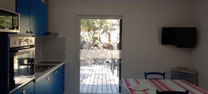 A kitchen with blue cabinets, a built-in oven, and a window overlooking a patio with a wooden deck and rocks. - Gîte Appicciu – 900m from the Beach (Coti-Chiavari)