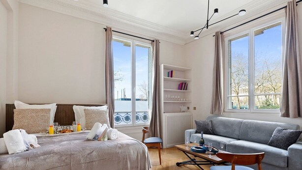 Ein Hotelzimmer mit Bett, einer Couch, einem kleinen Tisch, einem Bücherregal und großen Fenstern.