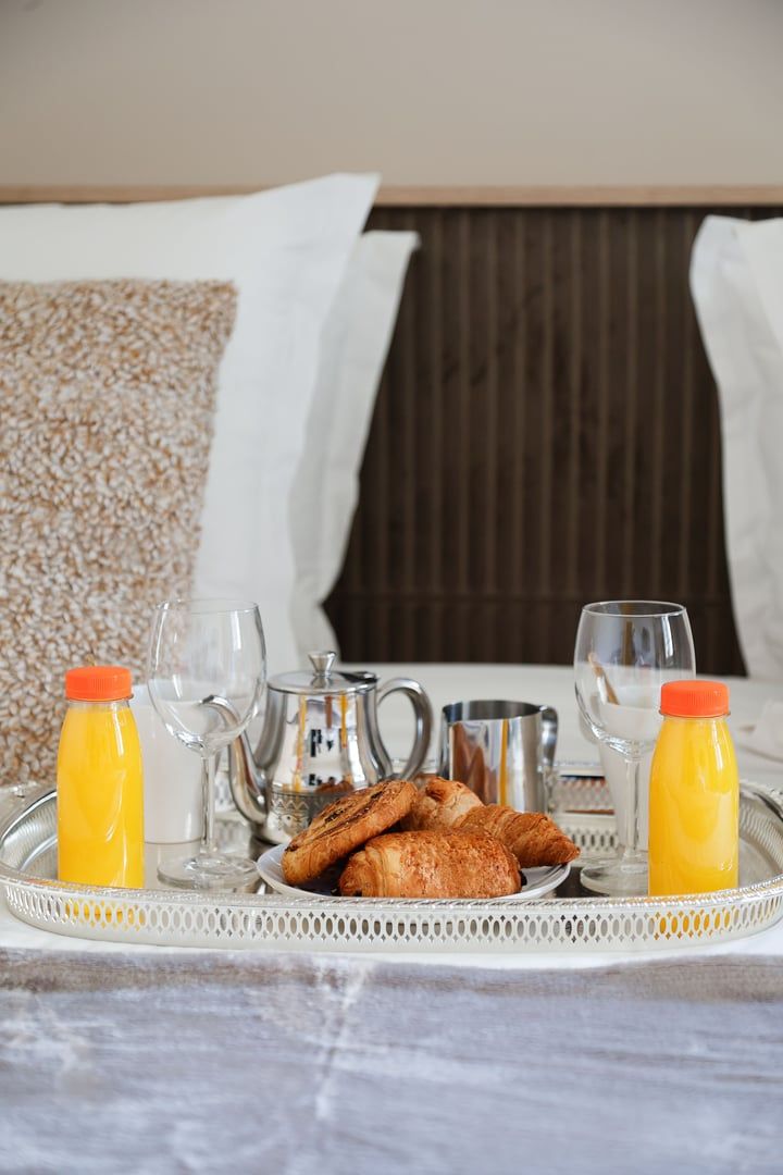 A breakfast tray with croissants, juice, and glasses on a bed with pillows and a textured wall.