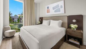 Een hotelkamer met een groot bed, een nachtkastje met een bloevas, een stoel en een raam met uitzicht op palmbomen.