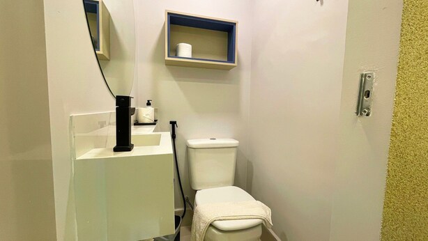 A small bathroom with a toilet, a sink with a soap dispenser, and a towel hanging on the wall. - Studio Vista Mar Lateral 6 - By Almare (João Pessoa)