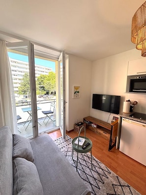 Un appartement moderne comprenant un canapé, un téléviseur, un coin cuisine et un balcon aménagé avec du mobilier d’extérieur.
