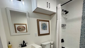 Een moderne badkamer met een wit wastafel, toilet en bad. Er hangen foto's in lijsten aan de muur, een handdoekhouder en een douchegordijn.