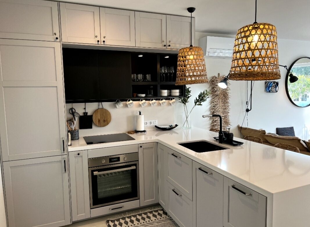 Una cocina moderna con armarios blancos, horno empotrado, un lavabo y dos lámparas colgantes de mimbre.