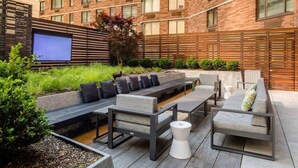 Een terras buiten met zitplaatsen, een televisiescherm en een bakstenen gebouw op de achtergrond.