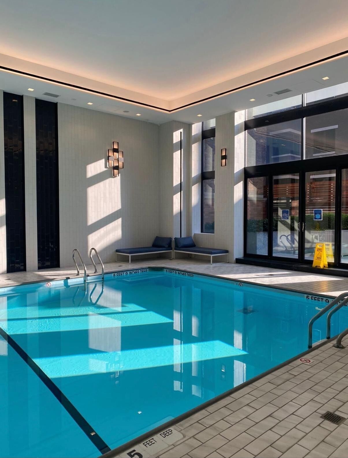 An indoor swimming pool with clear water, a pool ladder, and a bench area.
