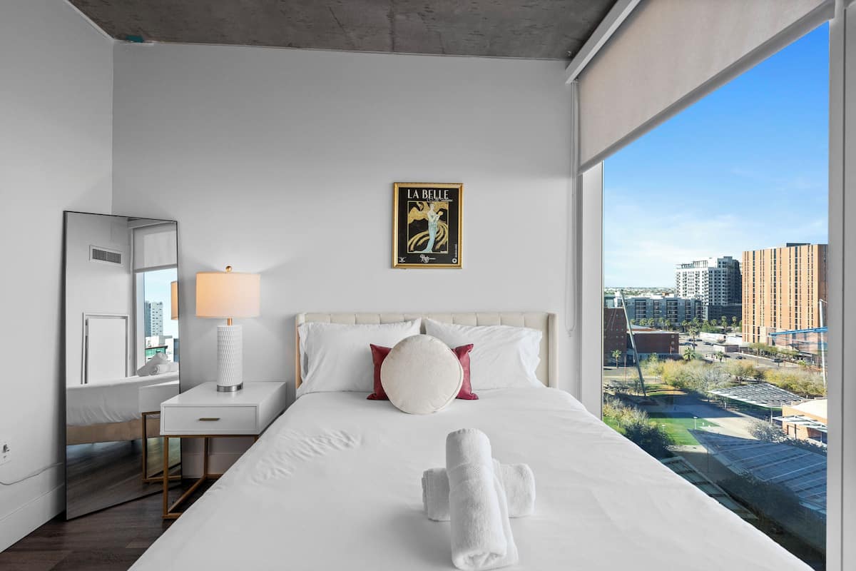 Habitación de hotel con una cama grande, una mesita de noche con lámpara, un espejo y vistas a la ciudad.