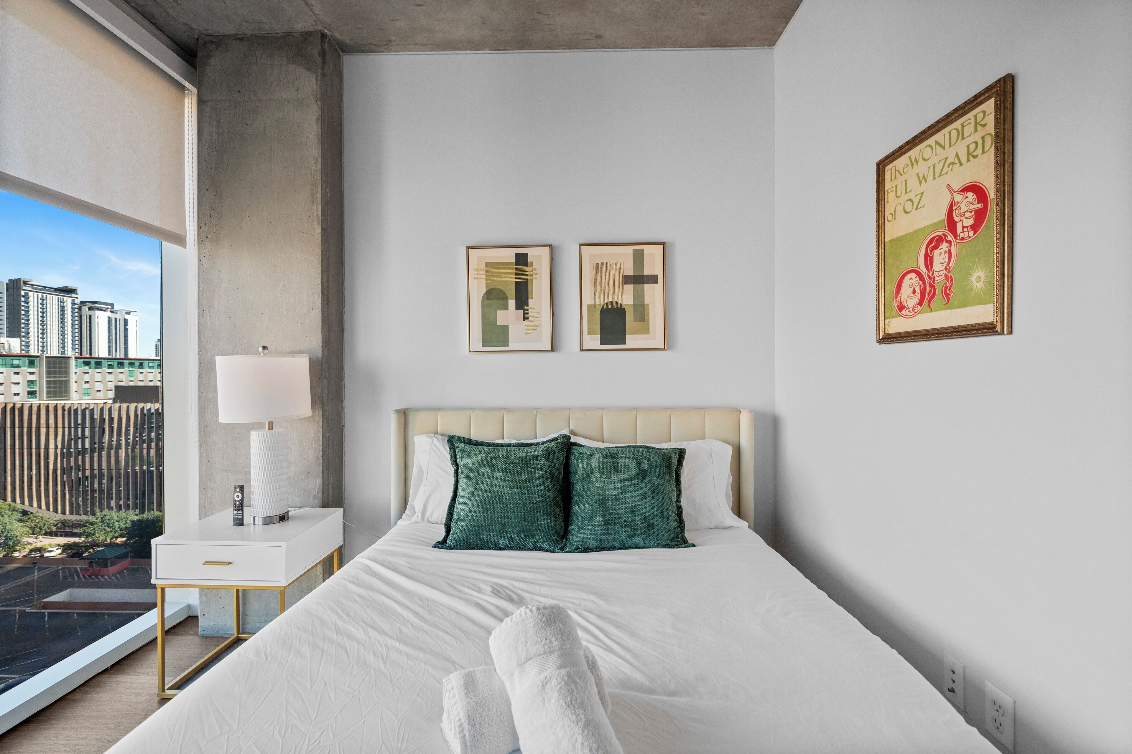 Habitación de hotel con cama, mesita de noche, lámpara, dos cuadros enmarcados y vistas a la ciudad.