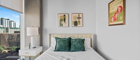 Habitación de hotel con cama, mesita de noche, lámpara, dos cuadros enmarcados y vistas a la ciudad.