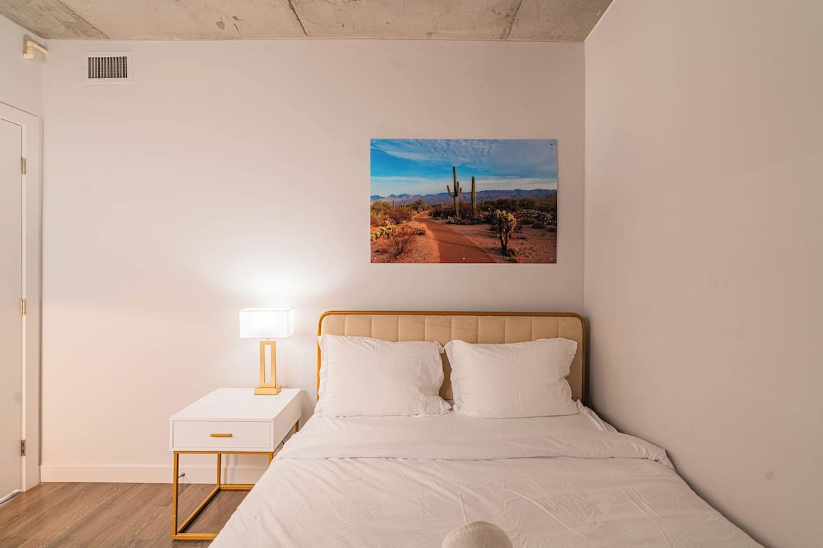 Un dormitorio con una cama, una mesita de noche con lámpara y un cuadro colgado en la pared que muestra un paisaje desértico.