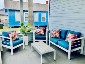 A covered outdoor seating area with white furniture and blue cushions, a small table, and a view of blue houses in the background. - Coastal Cove-Near Beach with a Game room (Galveston)