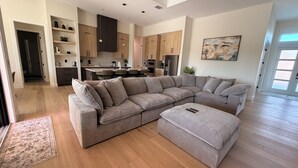 A modern living room with a large sectional sofa, a small ottoman, and a dining area with a kitchen in the background.