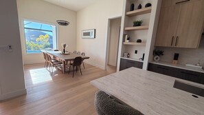 Moderni keittiö, jossa on puiset kaapit, ruokailutila pitkällä pöydällä ja tuoleilla sekä suuri ikkuna, josta avautuu näkymä ulos.