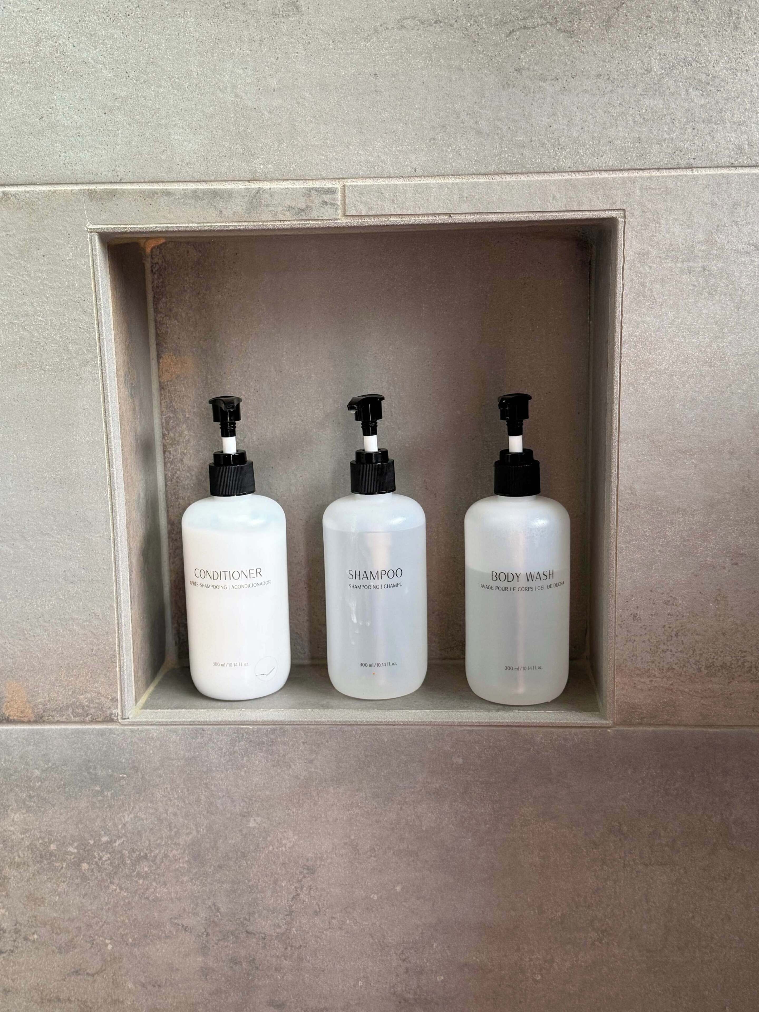 Drie flessen douchegel, conditioner en shampoo op een betegeld plankje.