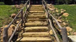 Una escalera de piedra conduce a un edificio de color oscuro con múltiples ventanas, rodeado de césped y rocas.