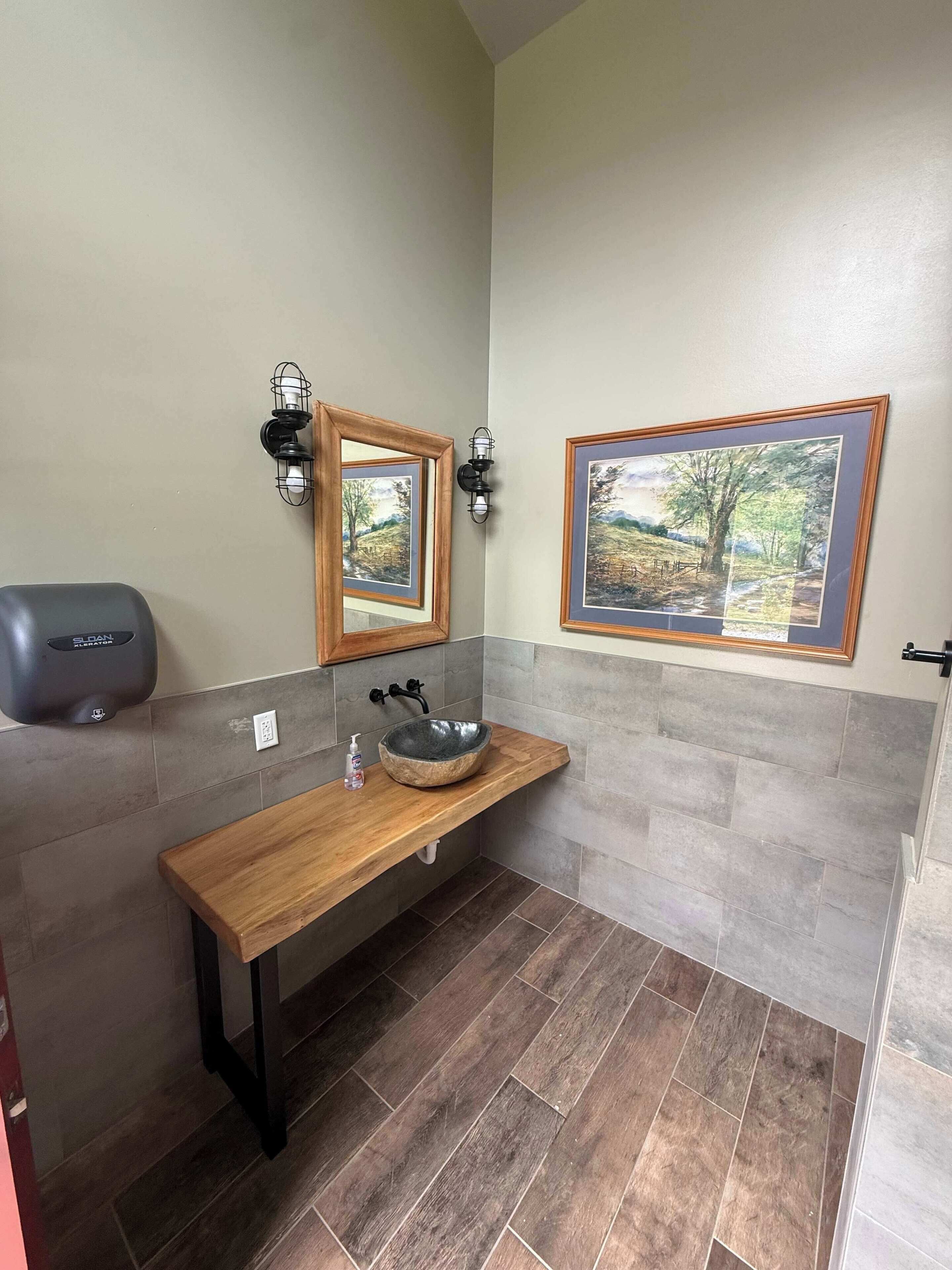 A bathroom with a wooden vanity, a stone sink, a wall-mounted mirror, two framed landscape paintings, and a hand sanitizer dispenser.