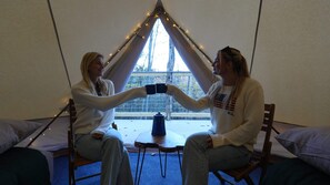 Two people seated in a tent-like room with a table and chairs, a window with curtains, and string lights.