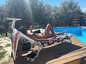 Eine Person liegt auf einer Liege am Pool, mit einem hölzernen Deck und blauem Himmel im Hintergrund.