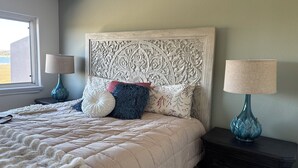 Ein Schlafzimmer mit einem großen Bett, einem dekorativen Kopfteil, zwei Nachttischlampen und Blick ins Freie durch ein Fenster.