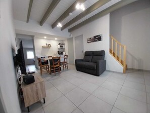 A modern living room with a sofa, dining table, chairs, a TV, and a staircase. - La Caillère Vacation Rental for 4 People (Moncoutant-sur-Sèvre)