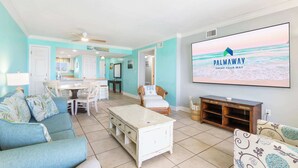 Een woonkamer met een tv waarop 'PALMAWAY VACAY YOUR WAY' staat, een eethoek met een tafel en stoelen, en een keuken op de achtergrond.